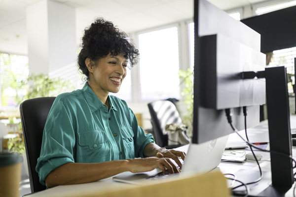 a smiling woman in a bright green shirt sitting at a desk and typing on a notebook computer