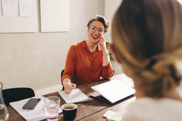 a woman sitting at a desk, smiling, with another woman, back to camera, facing her